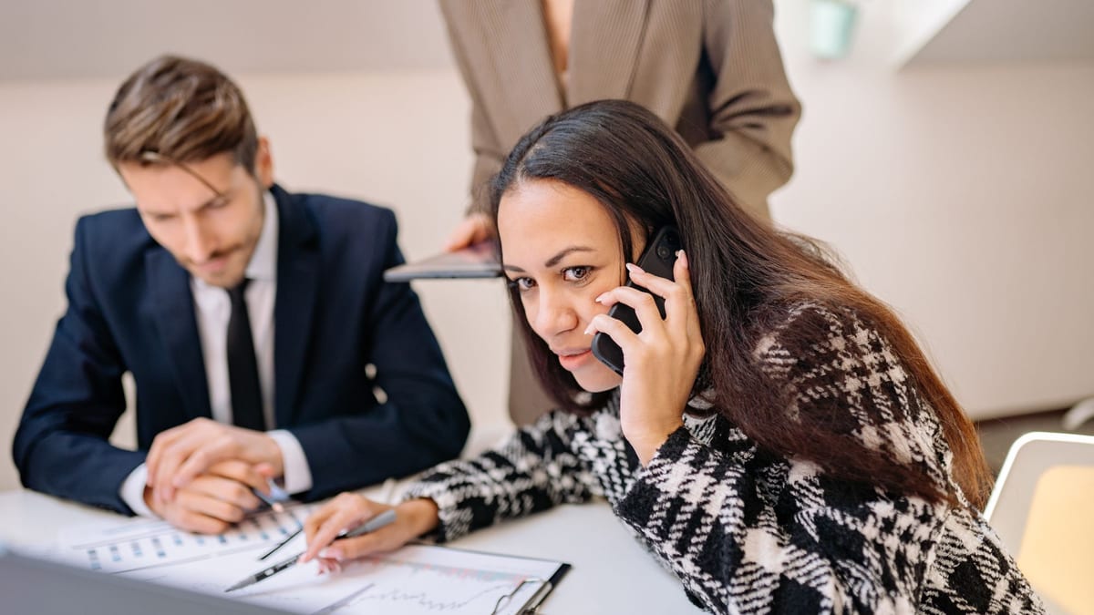 A woman on a phone call at a desk while a colleague in a suit writes notes beside her