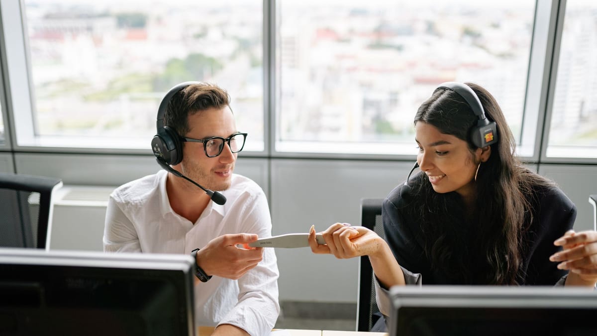 A man and a woman in headsets passing a document between them at their desks